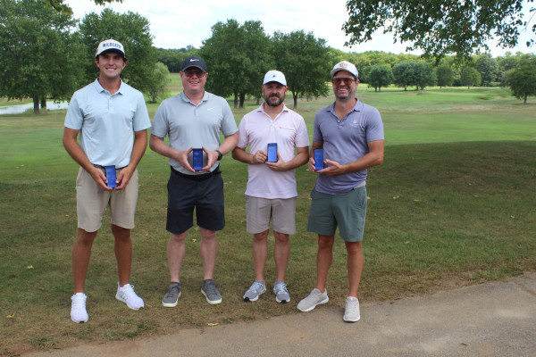 Four people stand outdoors on a golf course, each holding a blue rectangular award and smiling toward the camera. Trees and green fairways are visible in the background.