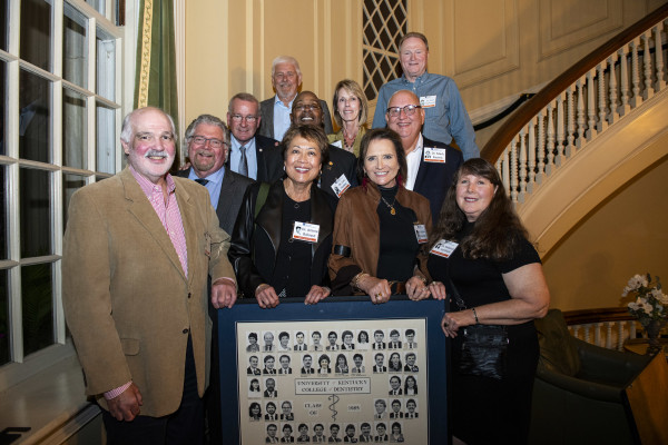 A group of alumni from the University of Kentucky College of Dentistry Class of 1985 poses together inside a warmly lit room with elegant architectural details. They stand on and around a staircase, smiling at the camera while holding a framed class composite featuring their graduation photos.
