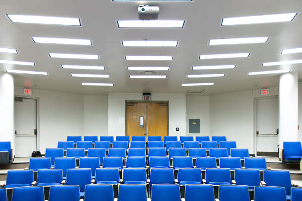 A brightly lit lecture hall with rows of empty blue chairs facing the front of the room. The ceiling is lined with rectangular fluorescent lights and a projector is mounted above the seats. At the front, double wooden doors are centered between two exit doors, all set against white walls.