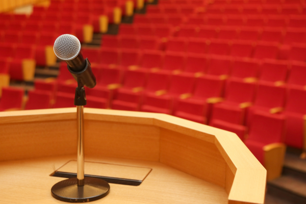 A close-up view from behind a wooden podium with a microphone positioned at the center. Beyond the podium is an empty auditorium filled with rows of red cushioned seats, fading into the background. The scene suggests a lecture, presentation, or event awaiting an audience.