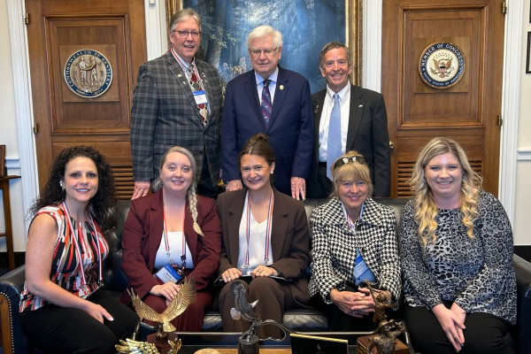 A group of eight individuals pose for a photo inside a government office, with three men standing behind five seated women; the room features wooden doors displaying the seals of the Commonwealth of Kentucky and a Member of Congress, and a large framed painting hangs behind them.