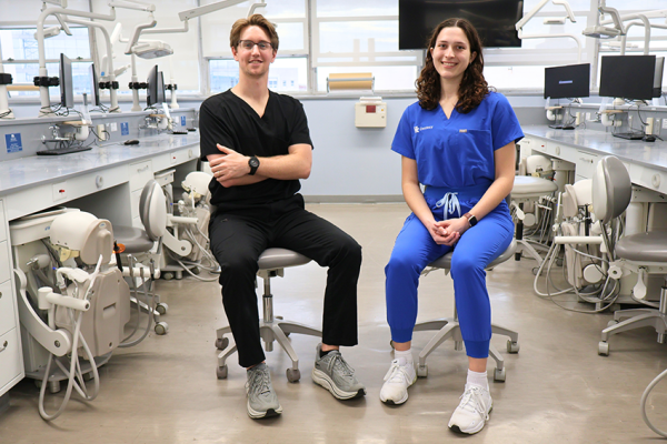 Two dental students sit on stools in a clinical simulation lab surrounded by dental chairs and equipment. One student wears black scrubs and the other wears blue scrubs with a university dentistry logo. Both are smiling toward the camera in a bright training clinic setting.