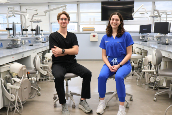 Two dental students sit on stools in a clinical simulation lab surrounded by dental chairs and equipment. One student wears black scrubs and the other wears blue scrubs with a university dentistry logo. Both are smiling toward the camera in a bright training clinic setting.