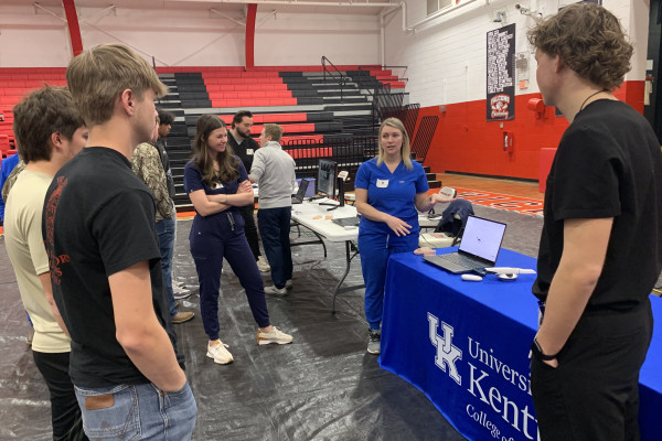 University of Kentucky College of Dentistry students and staff speak with high school students at an informational table inside a school gymnasium. A staff member in blue scrubs gestures while explaining materials displayed on a laptop, as several high school students stand nearby listening. The table is covered with a University of Kentucky College of Dentistry tablecloth, and additional demonstration stations and red-and-black bleachers are visible in the background.