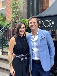 A smiling couple stands arm-in-arm outside a building. The woman wears a sleeveless black dress with a floral sash and gold accessories. The man wears a light blue blazer, white shirt, and dark pants. Behind them is a black awning with the word “FEDORA.”
