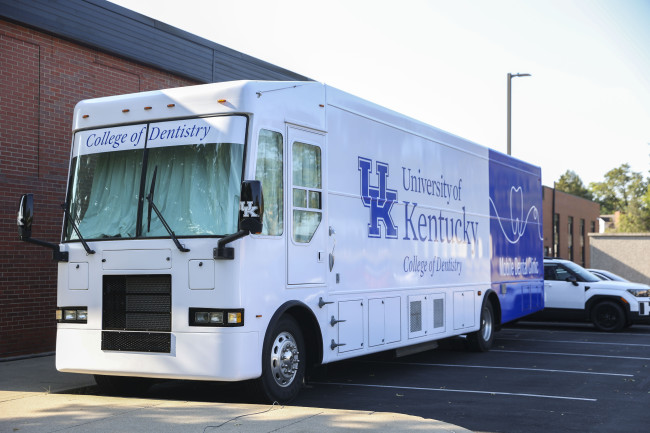 A large white and blue mobile dental clinic labeled “University of Kentucky College of Dentistry” is parked outside a brick building on a sunny day.