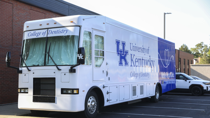 A large white and blue mobile dental clinic labeled “University of Kentucky College of Dentistry” is parked outside a brick building on a sunny day.