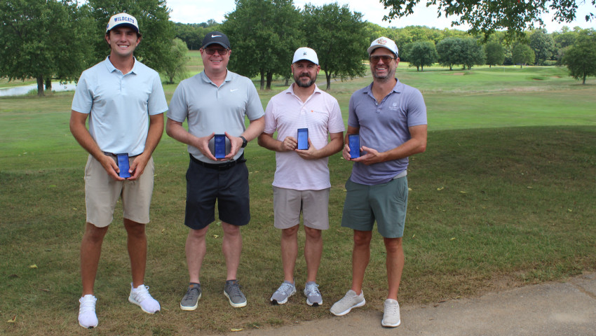 Four people stand outdoors on a golf course, each holding a blue rectangular award and smiling toward the camera. Trees and green fairways are visible in the background.