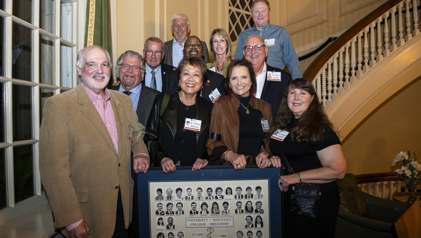 A group of alumni from the University of Kentucky College of Dentistry Class of 1985 poses together inside a warmly lit room with elegant architectural details. They stand on and around a staircase, smiling at the camera while holding a framed class composite featuring their graduation photos.