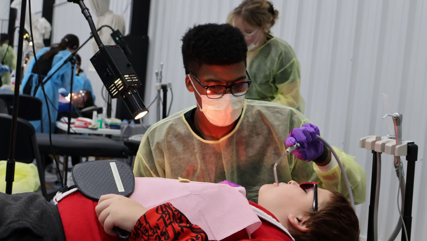 Dental student Donte Reed wearing protective gear and gloves provides care to a child reclining in a dental chair, using suction while other dental students work in the background of a clinic setting.