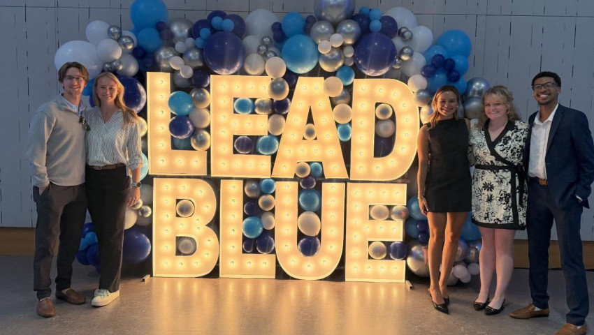Six University of Kentucky dental students stand smiling on either side of a large illuminated “LEAD BLUE” sign, decorated with blue, white, and silver balloons. The group is dressed in semi-formal attire, posing at the Lead Blue student awards event.