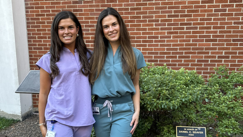 Twin sisters Kaylee and Kara Morris stand together outside the University of Kentucky College of Dentistry, smiling in front of a brick wall with large “College of Dentistry” lettering. One wears lavender scrubs and the other wears teal scrubs, 
