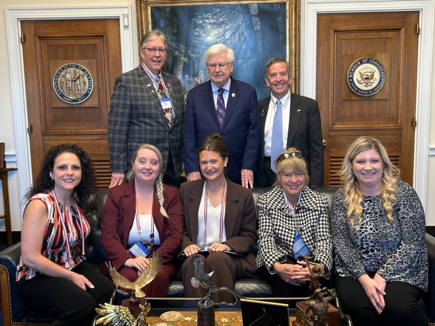 A group of eight individuals pose for a photo inside a government office, with three men standing behind five seated women; the room features wooden doors displaying the seals of the Commonwealth of Kentucky and a Member of Congress, and a large framed painting hangs behind them.