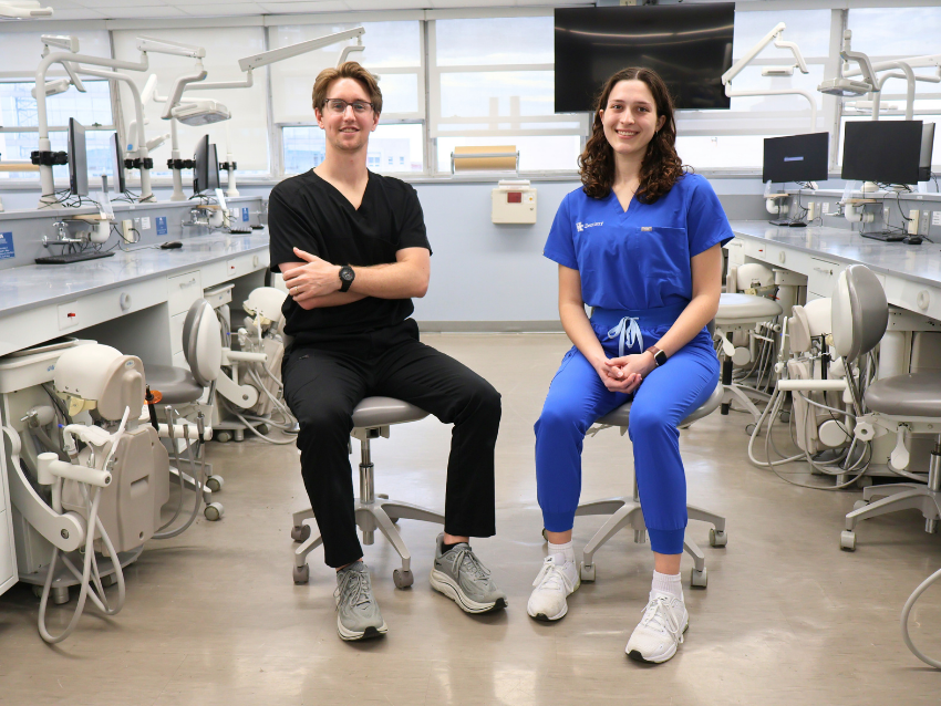 Two dental students sit on stools in a clinical simulation lab surrounded by dental chairs and equipment. One student wears black scrubs and the other wears blue scrubs with a university dentistry logo. Both are smiling toward the camera in a bright training clinic setting.