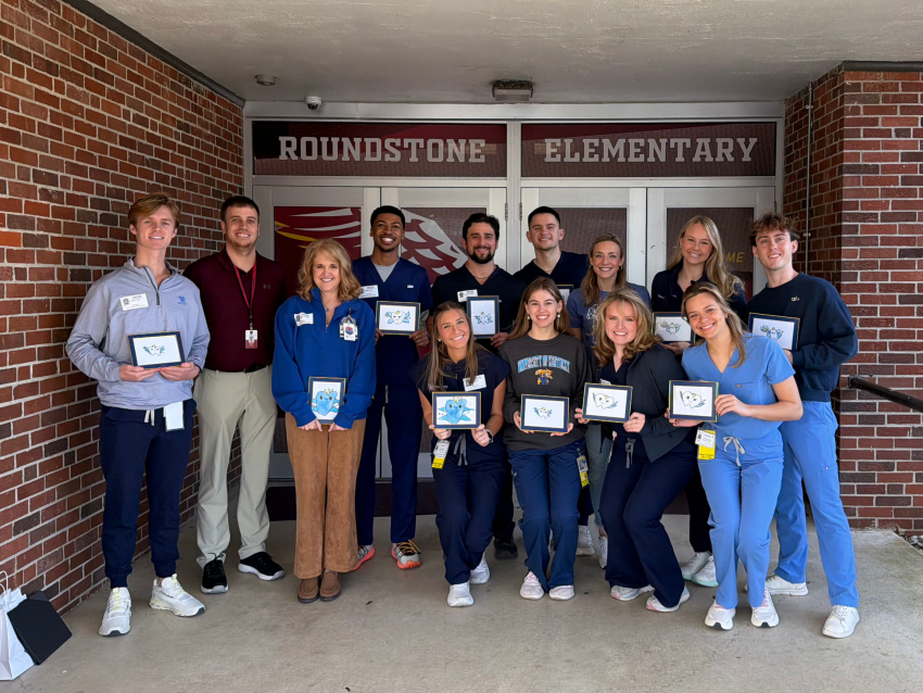 A group of University of Kentucky College of Dentistry students and faculty stand outside the entrance of Roundstone Elementary School, smiling and holding small framed drawings. The group is gathered beneath a covered entryway with brick walls and a sign reading “Roundstone Elementary” above the doors.