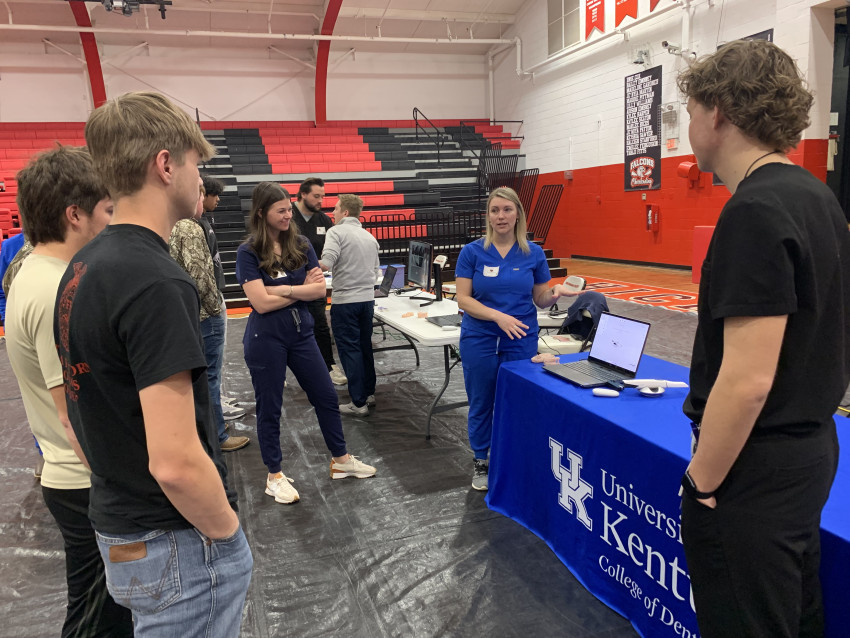 University of Kentucky College of Dentistry students and staff speak with high school students at an informational table inside a school gymnasium. A staff member in blue scrubs gestures while explaining materials displayed on a laptop, as several high school students stand nearby listening. The table is covered with a University of Kentucky College of Dentistry tablecloth, and additional demonstration stations and red-and-black bleachers are visible in the background.
