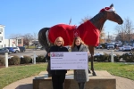 Two female UK representatives stand outdoors holding an oversized check in front of a bronze horse statue dressed in a holiday hat and red blanket. The check, dated December 9, 2025, awards $35,976 to the University of Kentucky College of Dentistry for Dental Assistant/EDDA training.