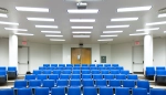 A brightly lit lecture hall with rows of empty blue chairs facing the front of the room. The ceiling is lined with rectangular fluorescent lights and a projector is mounted above the seats. At the front, double wooden doors are centered between two exit doors, all set against white walls.