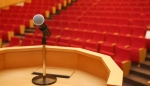 A close-up view from behind a wooden podium with a microphone positioned at the center. Beyond the podium is an empty auditorium filled with rows of red cushioned seats, fading into the background. The scene suggests a lecture, presentation, or event awaiting an audience.
