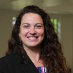 Portrait of Dr. Luciana Shaddox, who has long curly brown hair and is smiling. She is wearing a black blazer with a colorful top and a science-themed pin. The background is softly blurred.