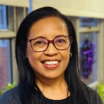 The image shows a woman smiling and wearing glasses, a dark top, and a gray cardigan. She is standing indoors near a window with soft natural light and some blurred background decorations.