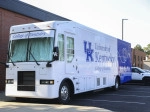 A large white and blue mobile dental clinic labeled “University of Kentucky College of Dentistry” is parked outside a brick building on a sunny day.