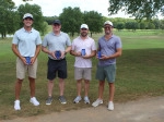 Four people stand outdoors on a golf course, each holding a blue rectangular award and smiling toward the camera. Trees and green fairways are visible in the background.