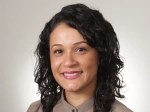 A woman with curly dark hair smiles at the camera, wearing a taupe blouse with a scalloped trim detail, posed against a plain light background.