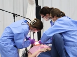 Three women in blue protective gowns, masks, and safety glasses work together to provide dental care to a patient lying back with a pink bib, as one operates equipment and another assists under bright task lighting.