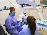 A dental provider wearing protective eyewear, gloves, and a blue mask performs a procedure on a patient using a large microscope while another team member assists chairside. Various dental instruments are organized on a nearby tray, and a computer workstation sits beside the operatory setup.