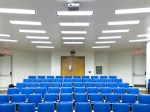 A brightly lit lecture hall with rows of empty blue chairs facing the front of the room. The ceiling is lined with rectangular fluorescent lights and a projector is mounted above the seats. At the front, double wooden doors are centered between two exit doors, all set against white walls.