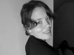 Black-and-white portrait of a person smiling over their shoulder, with short wavy hair and a dark top, posed against a plain indoor background.
