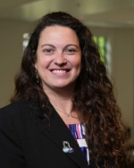 Portrait of Dr. Luciana Shaddox, who has long curly brown hair and is smiling. She is wearing a black blazer with a colorful top and a science-themed pin. The background is softly blurred.