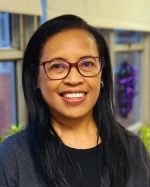 The image shows a woman smiling and wearing glasses, a dark top, and a gray cardigan. She is standing indoors near a window with soft natural light and some blurred background decorations.