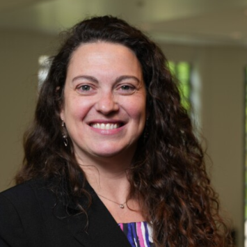 Portrait of Dr. Luciana Shaddox, who has long curly brown hair and is smiling. She is wearing a black blazer with a colorful top and a science-themed pin. The background is softly blurred.