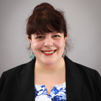 A woman with dark hair pulled back, wearing a black blazer over a white and blue floral top, smiling in front of a neutral gray background.