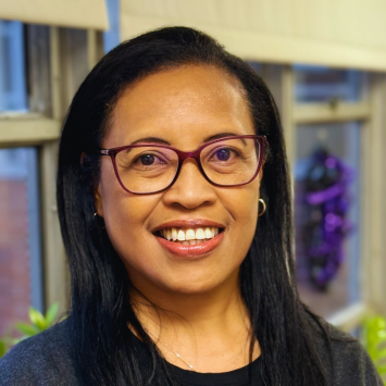 The image shows a woman smiling and wearing glasses, a dark top, and a gray cardigan. She is standing indoors near a window with soft natural light and some blurred background decorations.